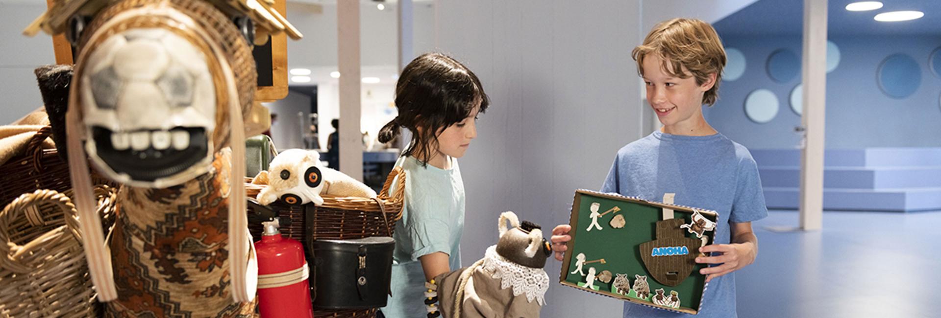 Zwei Kinder stehen einander zugewandt, eines mit einer Handpuppe und eines mit einem Diorama in der Hand.