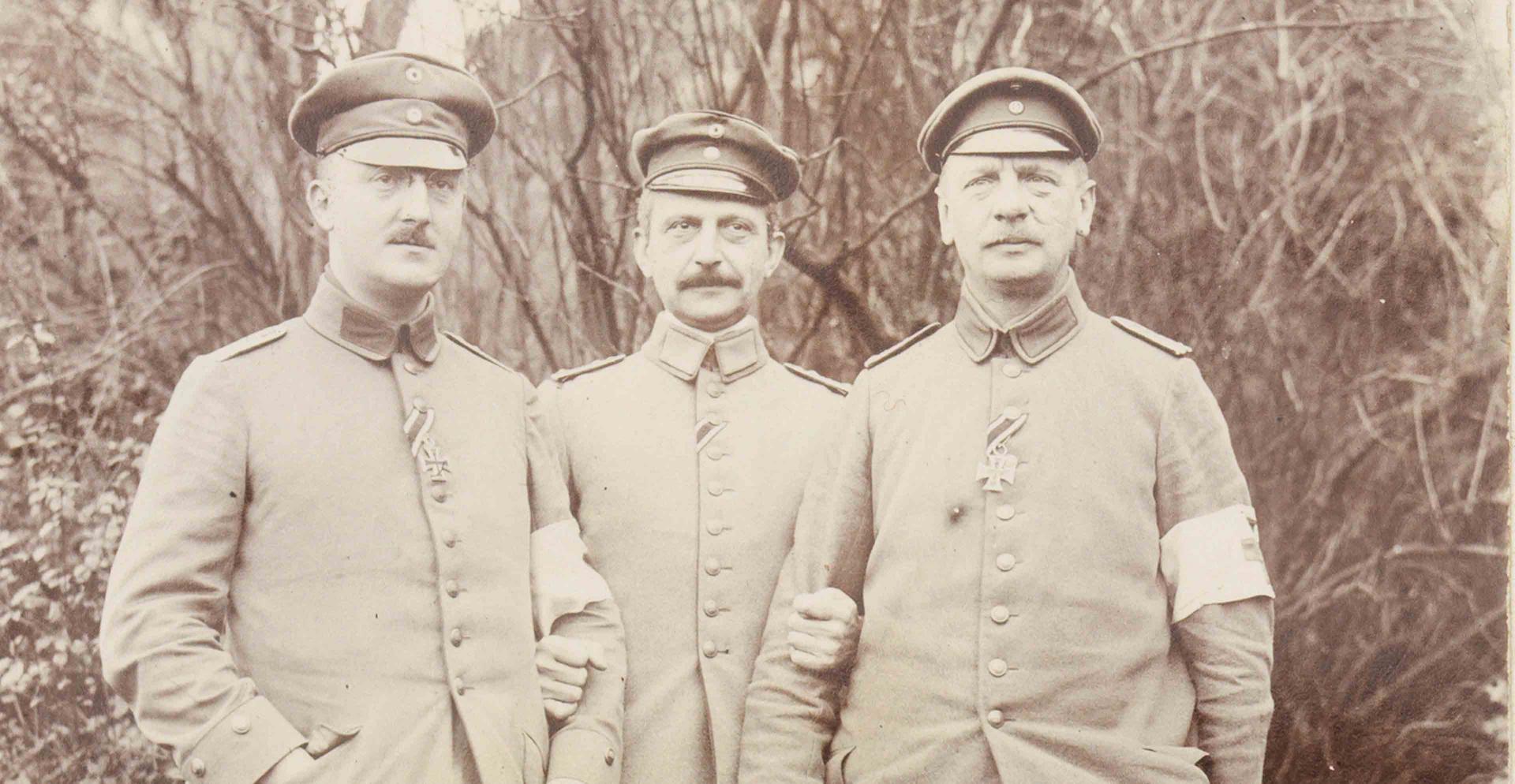 Schwarz-weiß Fotografie mit drei uniformierten Soldaten frontal und stehend vor einer Grünanlage.