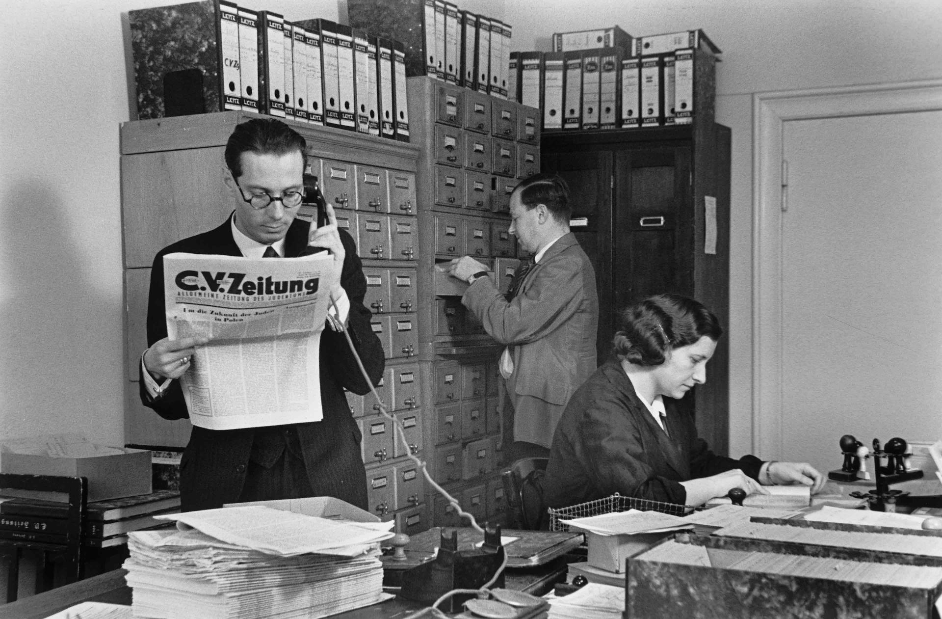 Ein Mann im Anzug liest im Stehen die C.V.-Zeitung und telefoniert dabei, im Hintergrund eine Frau am Schreibtisch und ein Mann am Aktenschrank
