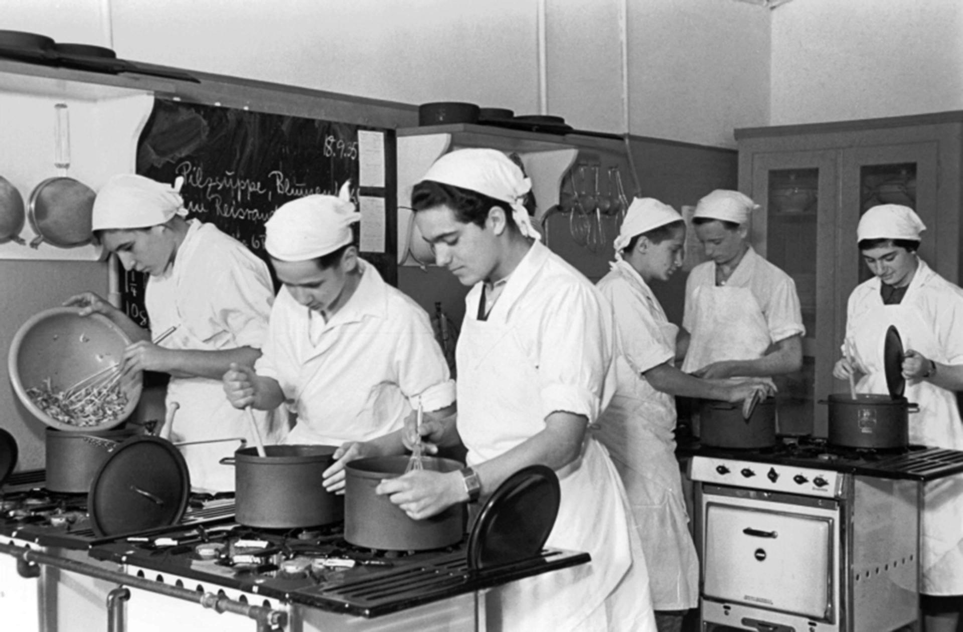 Black and white historical photograph of six students in a kitchen of the Theodor Herzl School, the students are wearing white aprons and white headscarves working hard at the stove cooking in pots