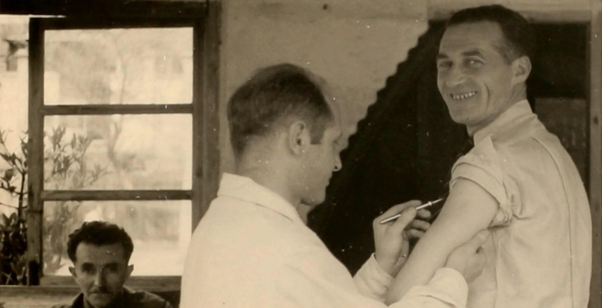 Black-and-white photograph of a man holding a scalpel to the upper arm of another man who is smiling for the camera