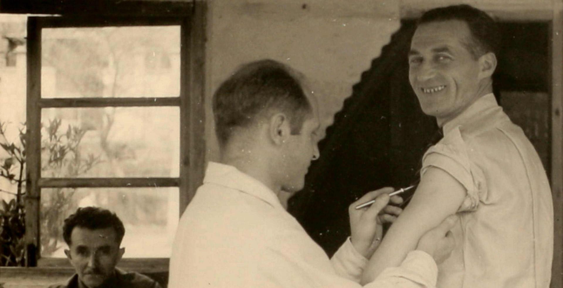 Schwarz-weiß-Foto eines Mannes, dem ein anderer Mann ein Skalpell an den Oberarm hält und der in die Kamera lächelt