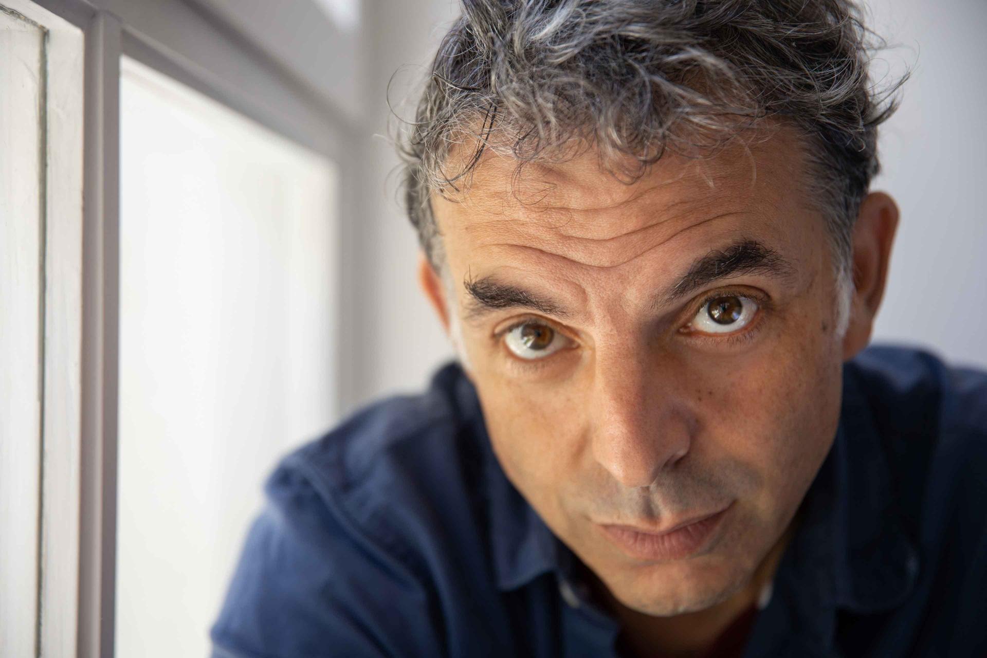 Portrait shot of a middle-aged man (Etgar Keret). His head is tilted slightly downward, he casts an intense gaze from diagonally below into the camera.