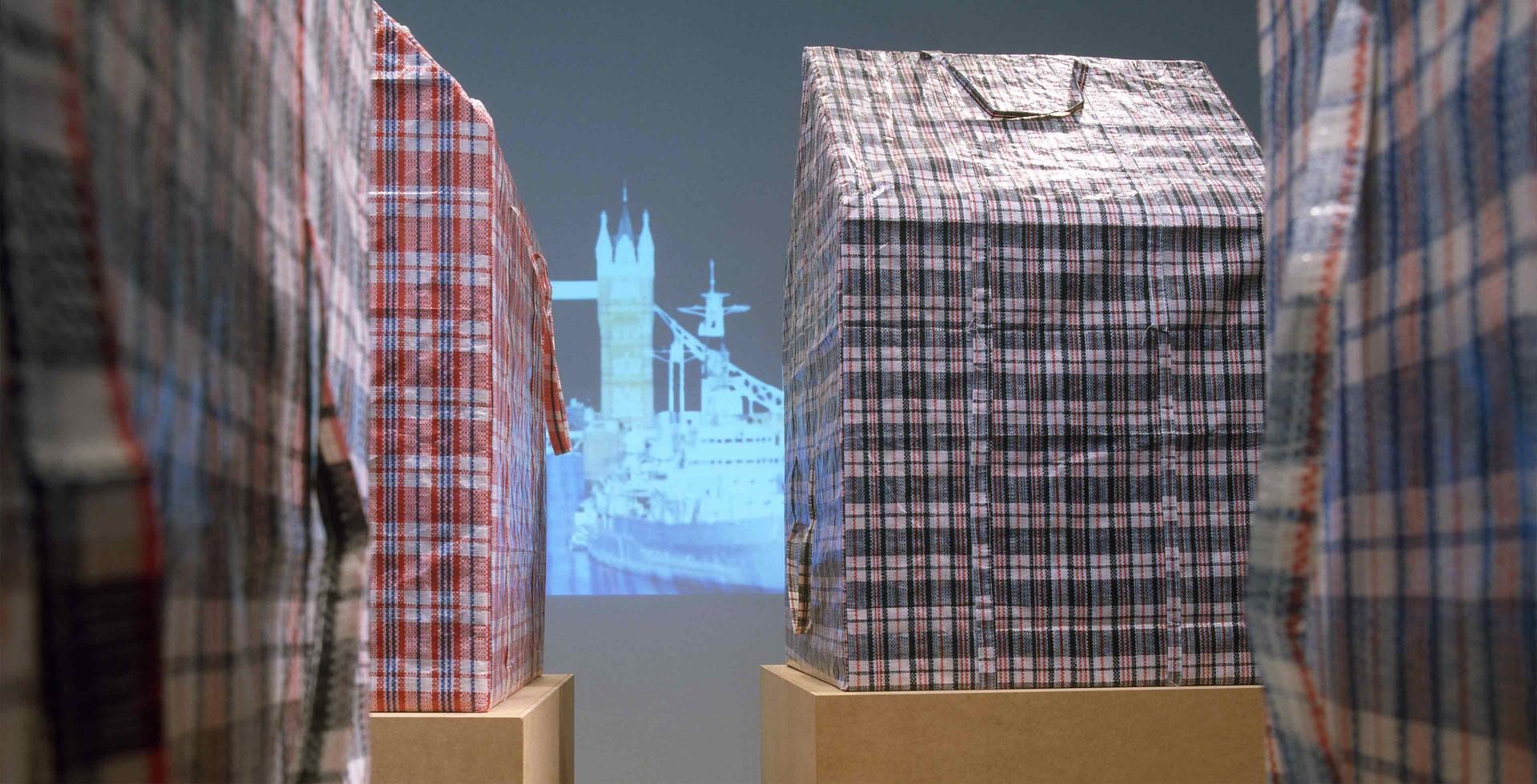 Houses made from red and white and blue checkered plastic travel bags, sitting on cardboard boxes