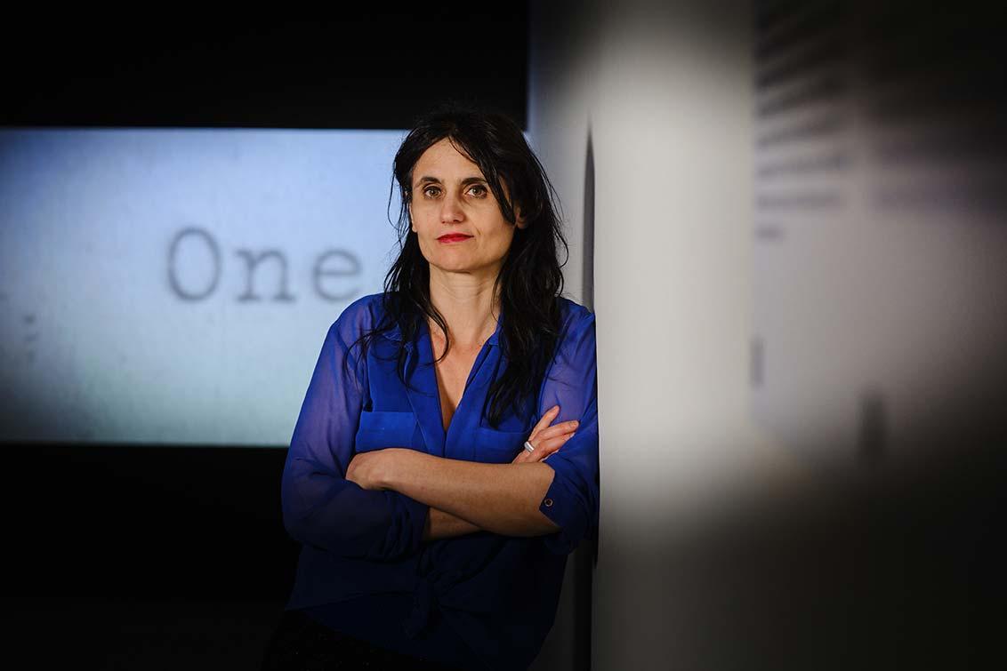 A woman with long dark hair leans against the wall in an exhibition room with her arms folded, behind her you can see a film screen on which the word "one" can be read.