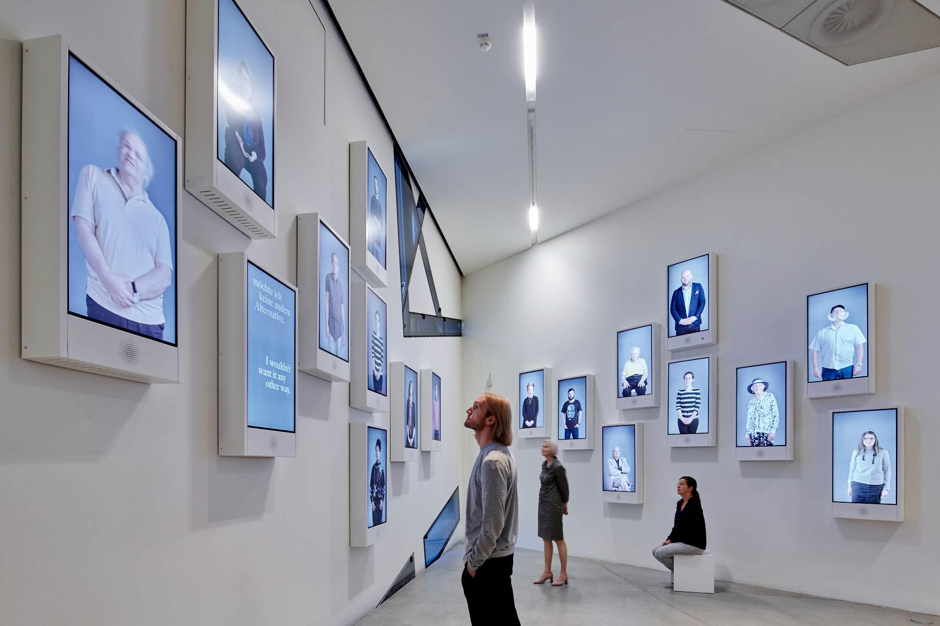 Visitors in an exhibition room with screens scattered along the walls showing videos of various people