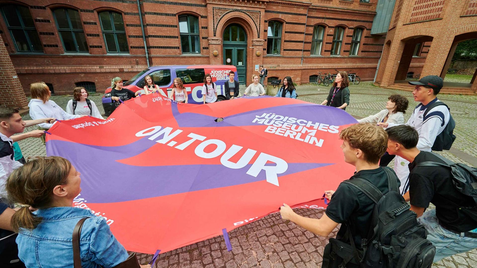 Eine Gruppe von Jugendlichen spannen auf einem Innenhof ein großes Tuch auf, auf dem on.tour und Jüdisches Museum Berlin steht.
