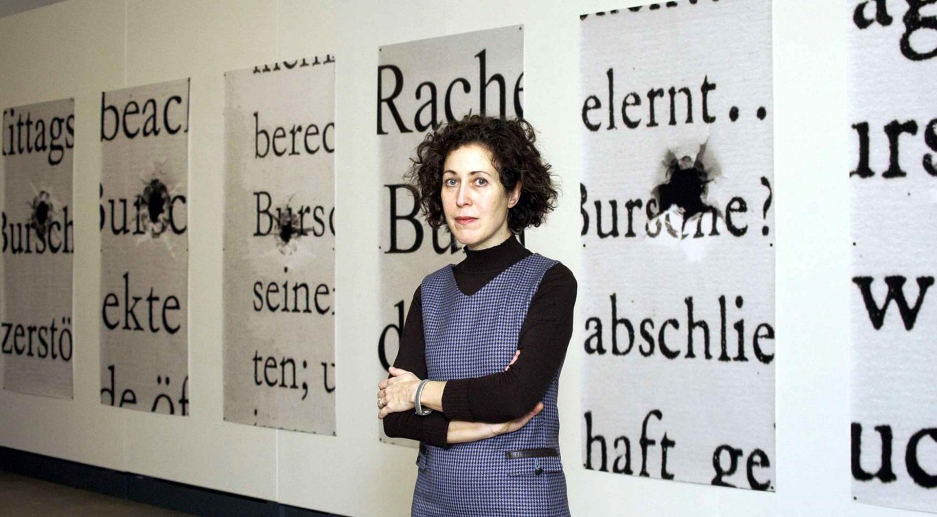 A woman stands in front of a wall on which posters with words in large letters and bullet holes can be seen