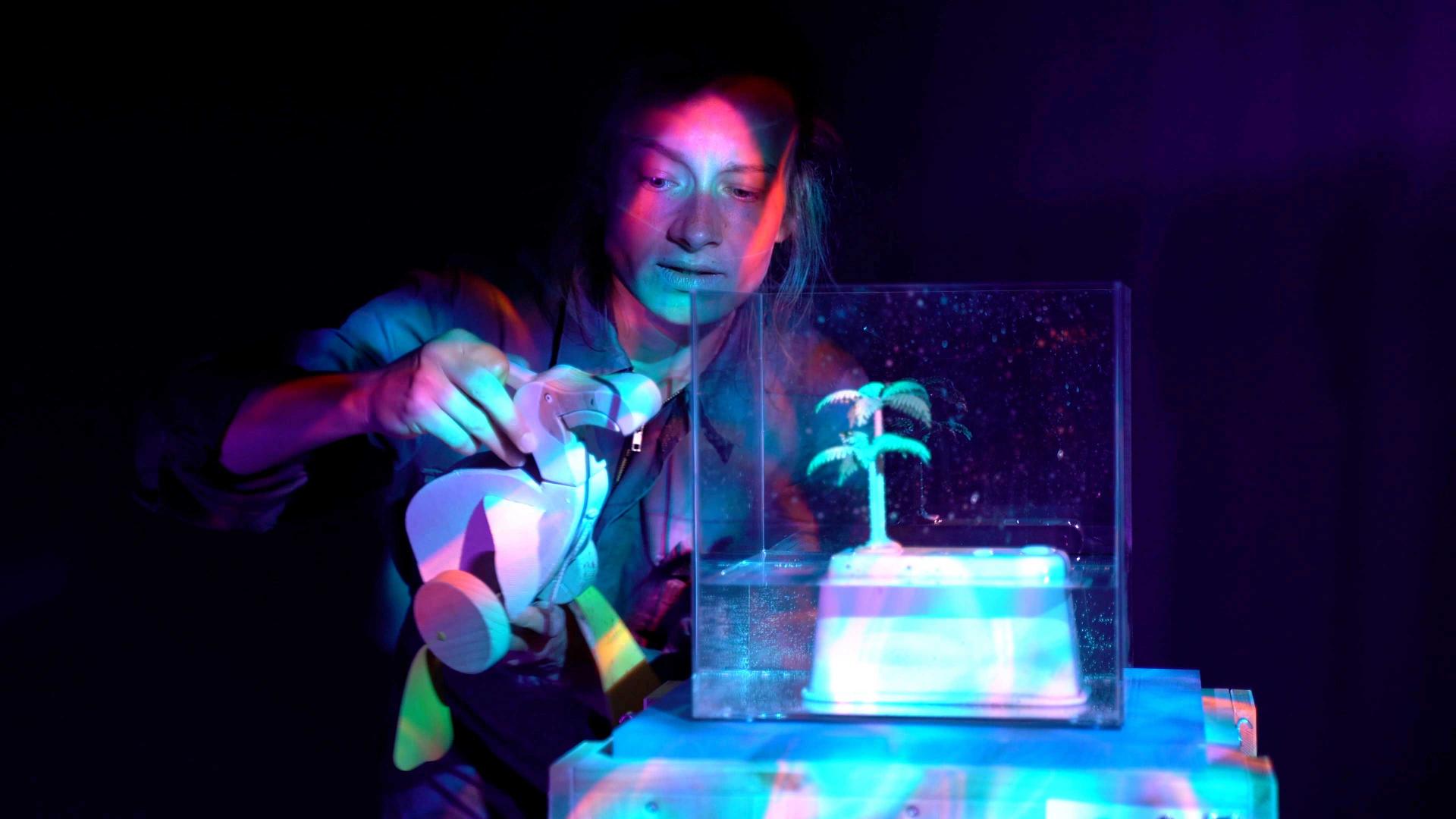 An actress holds a wooden toy in her hands and stands next to a white glass box containing a plastic palm tree. It is dark, the actress and the objects are illuminated by coloured light.