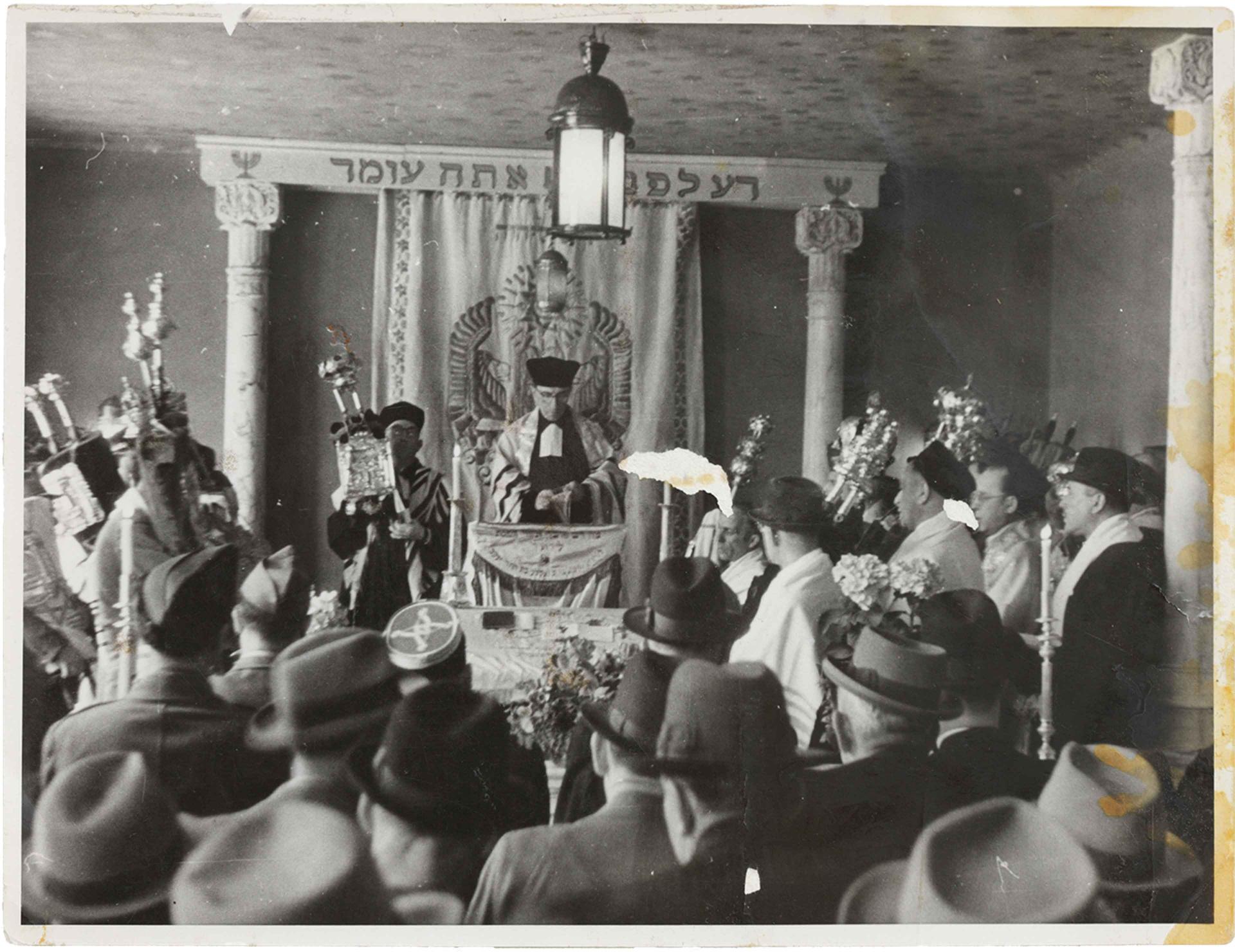 Black and white photograph of a synagogue, in the center a rabbi, around him men with Judaica 