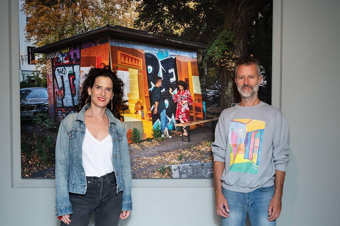 A woman and a man stand in front of a large-format photograph in the exhibition room, in which they can be seen in front of a brightly painted shed
