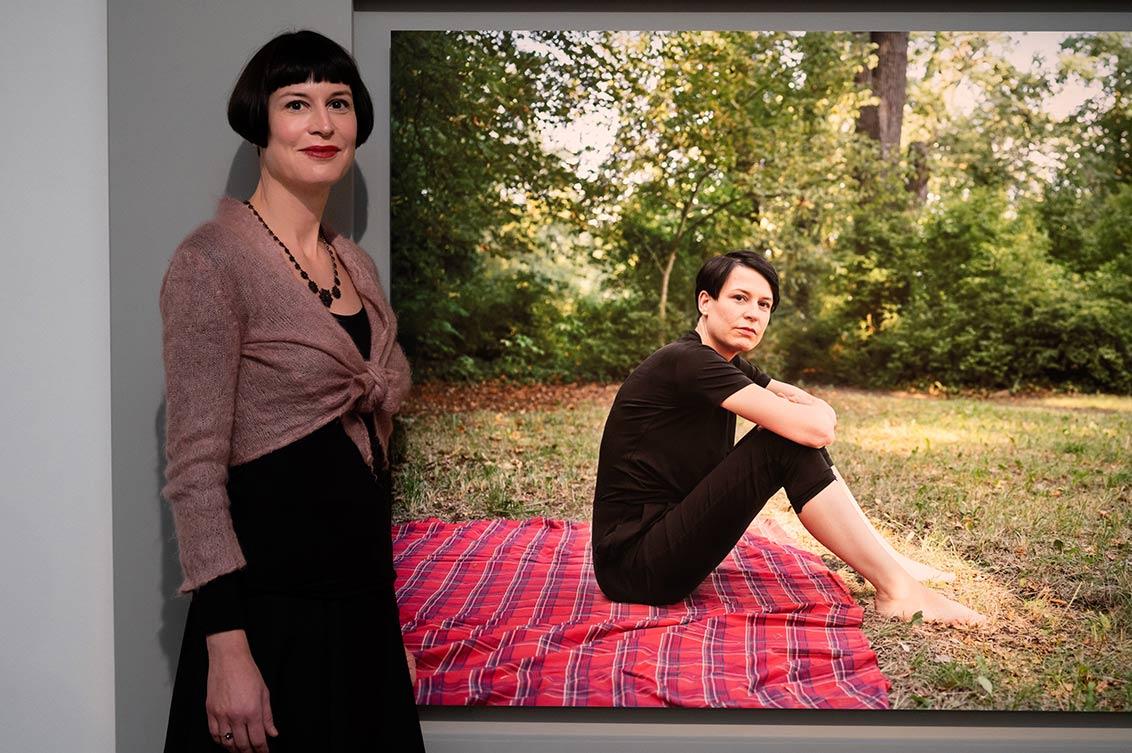 Barbara Steiner stands next to her large-format portrait in the exhibition, in the portrait she is sitting on a picnic blanket in a meadow