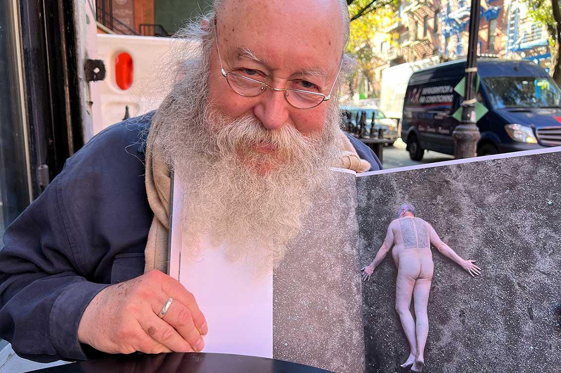 A man with a long white beard and glasses smilingly holds an open catalog with a photo of himself into the camera. He is sitting at a small café table, in the background a street with cars