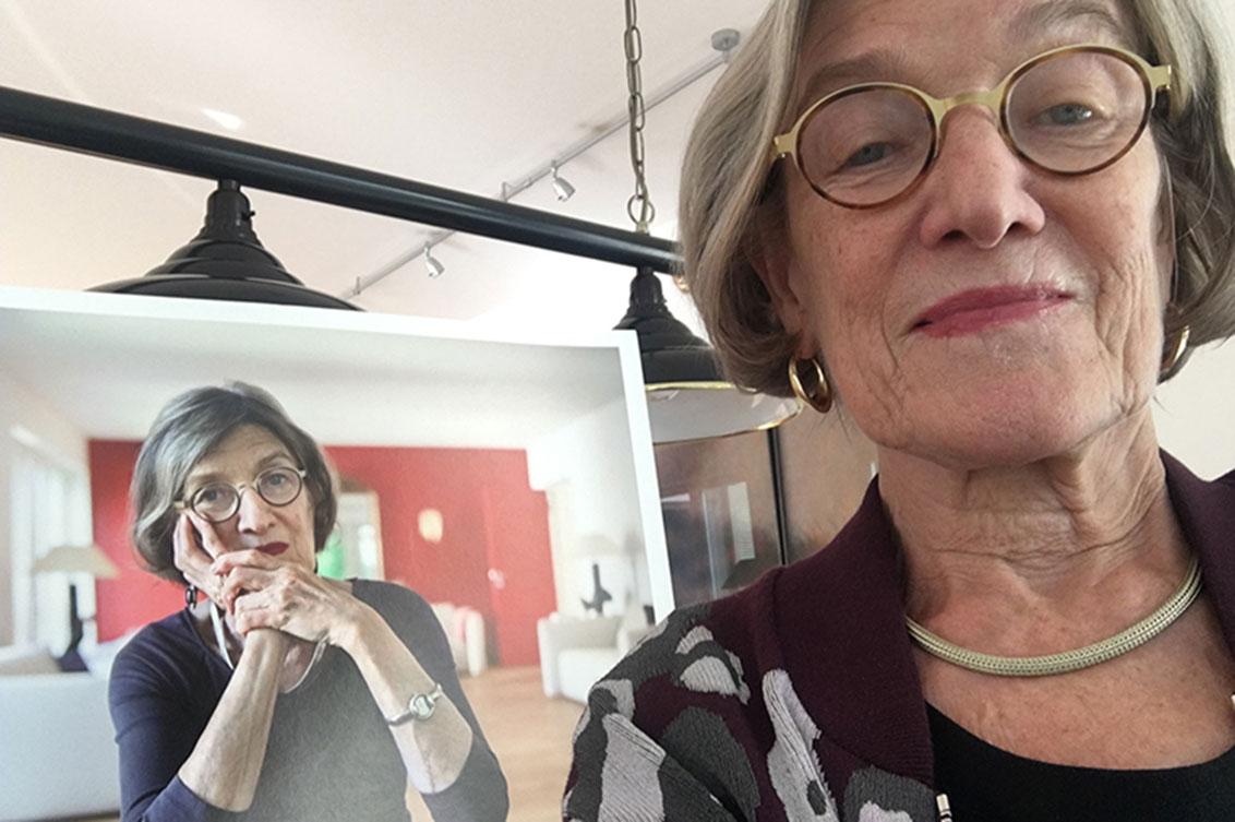 Selfie of woman with glasses in front of large print of her portrait, lamp is seen in background