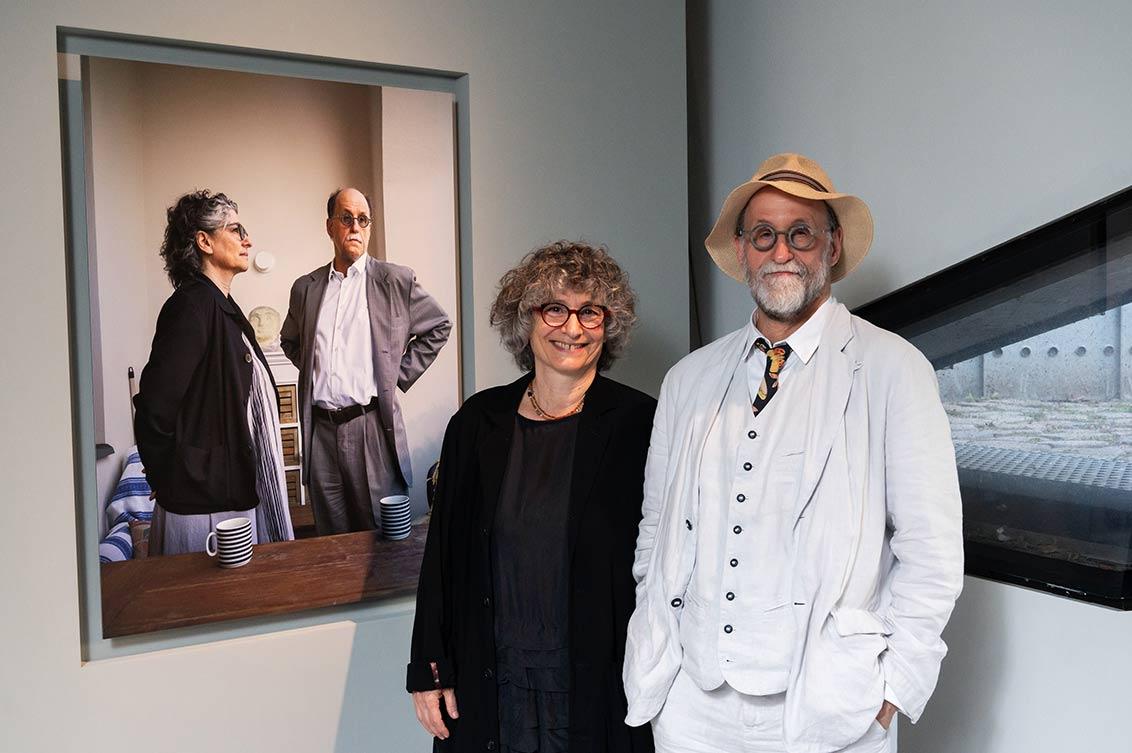 A smiling couple, both gray-haired, she all in black, he in a white suit and straw hat stands next to a portrait of them in the exhibition, in the portrait they are standing behind a table looking in different directions