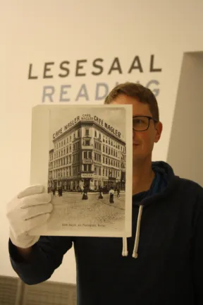 A man holding up a black and white photo in front of him. On the photo we see a bulding