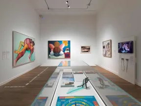Ausstellungsansicht, in der Mitte des Raums steht eine Glasvitrine, an den W&auml;nden h&auml;ngen farbenfrohe Bilder.