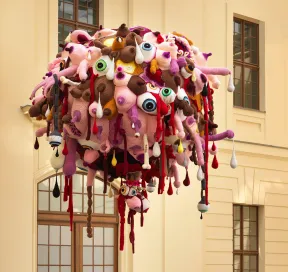 Ein gro&szlig;er Ball aus geh&auml;kelten K&ouml;rperteilen h&auml;ngt von der Decke des Glashofs. Der Ball besteht aus rosa, lila, braunen und wei&szlig;en geh&auml;kelten Augen, Penissen, Br&uuml;sten und Knochen.