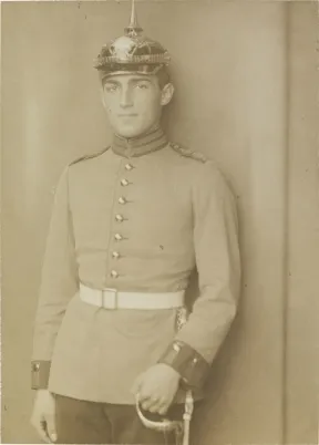 Black-and-white photograph: Young man in soldier&rsquo;s uniform with white belt, spiked helmet, and sword, studio portrait