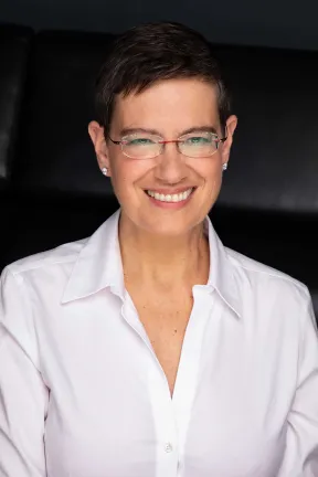 Eine Frau mit Brille und einer wei&szlig;en Bluse schaut freundlich in die Kamera.