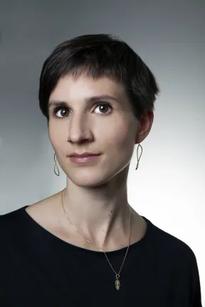 A woman with short hair looks cheerfully into the camera. She is wearing earrings and a necklace.