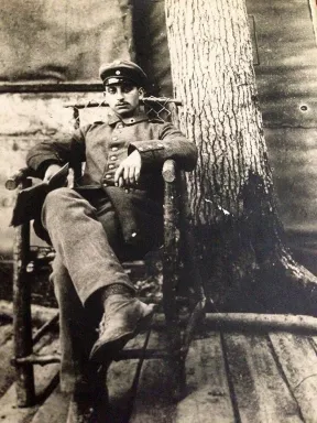 Schwarz-Wei&szlig;-Foto eines Mannes in Uniform, der auf einem Stuhl sitzt, die Arme auf den Lehnen, ein Bein &uuml;berschlagen, im Hintergrund ein Baumstamm, unter dem Stuhl ein Boden aus Holzdielen.