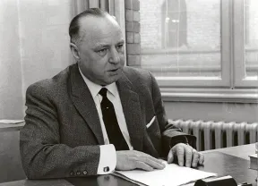 Black-and-white photo of a man wearing a suit and tie at a desk.