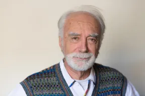 The photo shows an older man with white hair and beard looking directly into the camera. He is wearing a white shirt and a patterned slipover.