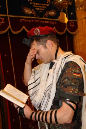 Ein Mann in Bundeswehruniform h&auml;lt in der linken Hand ein aufgeschlagenes Buch, die rechte Hand h&auml;lt er vor seine Augen, sein Blick ist gesenkt auf das Buch. An seinem linken Arm tr&auml;gt er einen j&uuml;dischen Gebetsriemen.