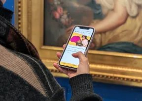 Das Bild zeigt links die Schulter sowie die Hand einer Person. Sie h&auml;lt ein Smartphone, auf der die App "Junge Perspektiven" ge&ouml;ffnet ist. Im Hintergrund sieht man den Ausschnitt eines Gem&auml;ldes.
