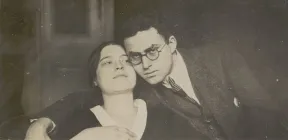 The historical photograph shows a young woman with a man leaning against her head on her right side. He is looking at the camera and has his right arm around the woman's shoulders.