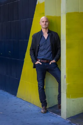 A man is standing in front of a yellow and blue wall. He has his hands in his trouser pockets.