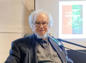 Farbbild. Ein &auml;lterer Herr sitzt auf einem Stuhl. Vor ihm steht ein Mikrofon. Er schaut direkt in die Kamera. 