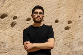Ein Mann mittleren Alters mit kurzen schwarzen Haaren, einer Brille und einem schwarzen T-Shirt. Der Mann hat die Arme verschr&auml;nkt und steht vor einer Felswand.