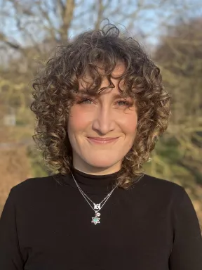 Portrait Foto einer l&auml;chelnden Frau. Sie tr&auml;gt einen schwarzen Pullover und eine Kette mit Davidstern Anh&auml;nger. Im Hintergrund ist unscharf ein Baum zu sehen.