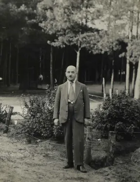 The image shows a man wearing a suit. He stand in front of bushes and trees.