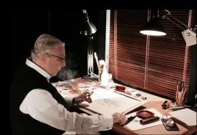 Ein Mann sitzt am Schreibtisch, raucht eine Zigarre und zeichnet etwas. 