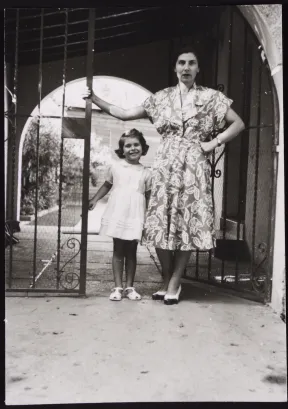 Black-and-white photo. A middle-aged woman and a little girl are standing next to a gate in front of a house entrance.