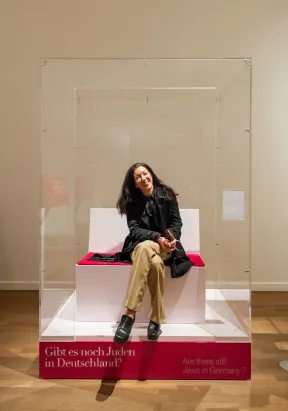 Sharon Adler sitzt auf einer Sitzbank in einer nach vorne hin offenen Plexiglas Box, auf dem Sockel steht: Gibt es noch Juden in Deutschland?