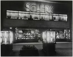 Schwarz-wei&szlig; Foto eines Geb&auml;udes mit leuchtenden Vitrinen und dem leuchtendem Schriftzug Scherk am Kurf&uuml;rstendamm bei Nacht.