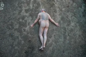 Photography of a naked man lying prone on gravelly earth, a passage from Adorno's Minima Moralia is tattooed in two columns on his back