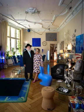 A couple, he in black suit with hat and white beard, she in floral dress with turquoise glasses, pose in a large, very colorfully decorated room, in front of them a blue bunny sculpture