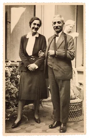 Schwarz-wei&szlig;-Fotografie zweier Menschen auf dem Vorplatz eines Hauses mit Blumen, Statue und Korbstuhl im Hintergrund