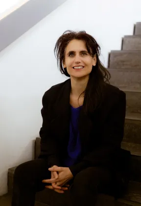 Eine Frau mit langen braunen Haaren, schwarzem Jackett und blauem Pullover sitzt auf einer Treppe und schaut l&auml;chelnd direkt in die Kamera.