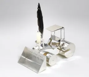 Farbfotografie einer realistisch gestalteten Miniatur-Dampfwalze mit Walzrolle, Baggerschaufel und F&uuml;hrerh&auml;uschen. Auf der Oberseite des stilisierten Motorraums sind ein Metallr&ouml;hrchen zum Einstecken einer Feder sowie ein Kerzenhalter angebracht.