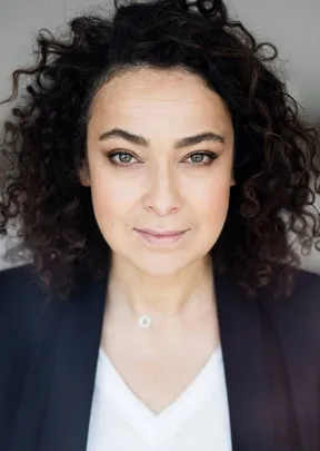 Portr&auml;tfoto einer Frau mit schwarzen Locken und blauem Blaser &uuml;ber wei&szlig;em Pulli, die direkt in die Kamera schaut.
