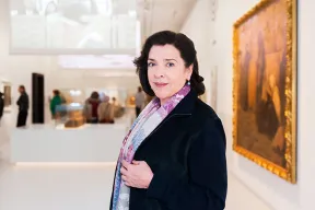 Auf dem Farbfoto ist Elene Bashkirova in schwarzem Blazer und violett gemustertem Schal zu sehen. Im Hintergrund ist die Jerusalem-Ausstellung des J&uuml;dischen Museums Berlin zu erkennen.