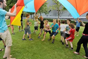 Kinder und Betreuer*innen spielen im Garten mit einem Schwungtuch