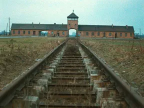 Bahngleise, die auf ein breitgestrecktes, flaches Backsteingeb&auml;ude zuf&uuml;hren. In der Geb&auml;udemitte befindet sich ein Wachturm mit einem Tor f&uuml;r die Durchfahrt ankommender Z&uuml;ge. Es handelt sich um das Lagertor des Vernichtungslagers Auschwitz-Birkenau.