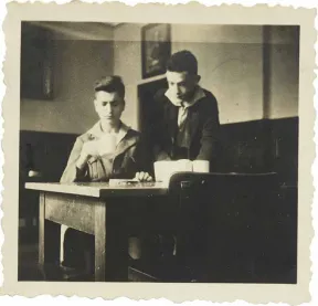 Two boys at a table, one sitting with a cup, one standing diagonally behind him (black-and-white photo).